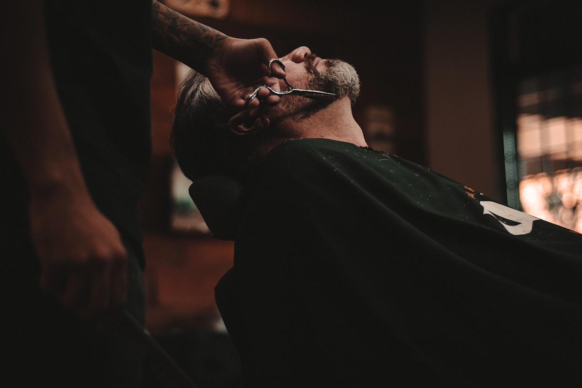 A barber trimming a reclining client’s beard