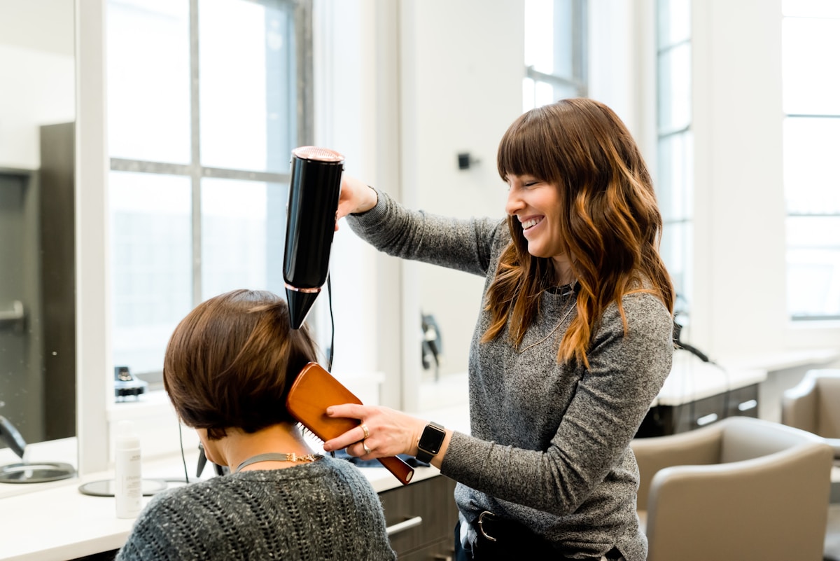 A stylist blow-drying a seated client in a sunlit salon